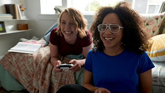 Two smiling women playing Xbox together in a bedroom with one woman sitting on the floor against the bed and the other laying on the bed while playing