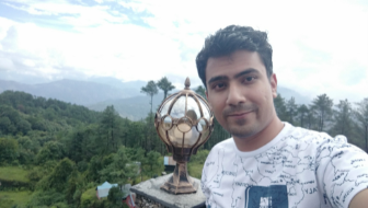 KnightlySpartan takes a selfie with forest-covered mountains or hilltops in the background. Also in the background, a metal and glass item on a stone pedestal. His reflection is showing in the glass. 